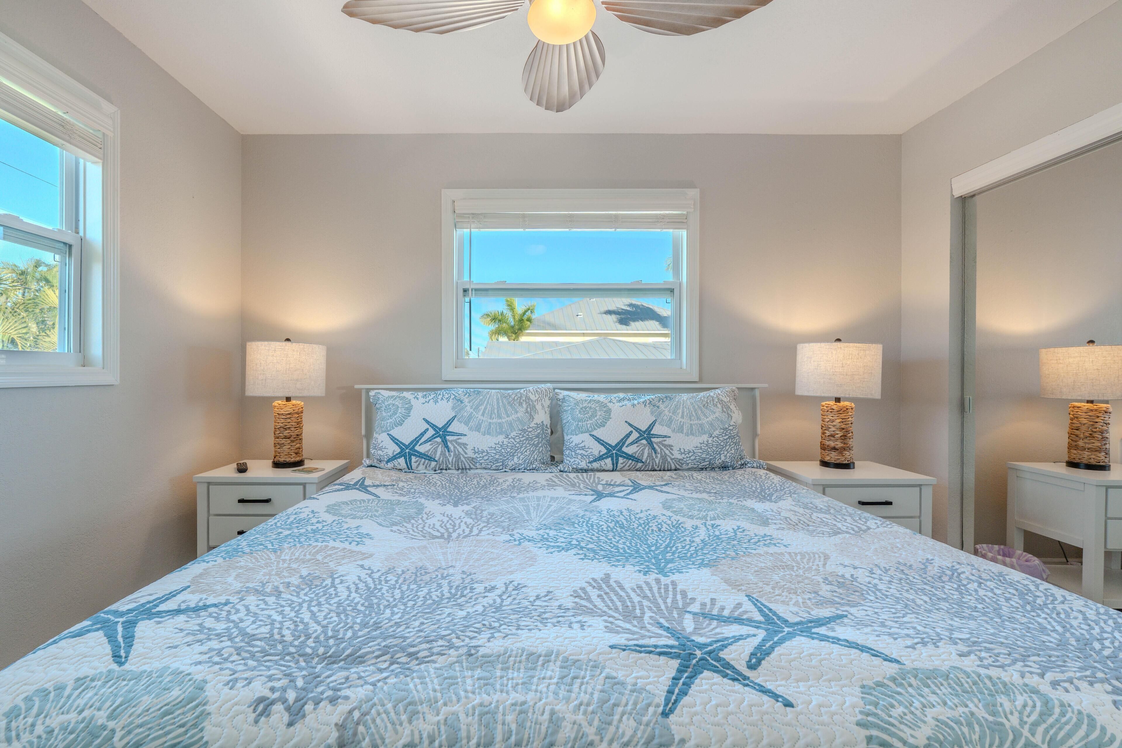 A bedroom with a bed, two nightstands, two lamps, a window with a view of the beach, and a ceiling fan.