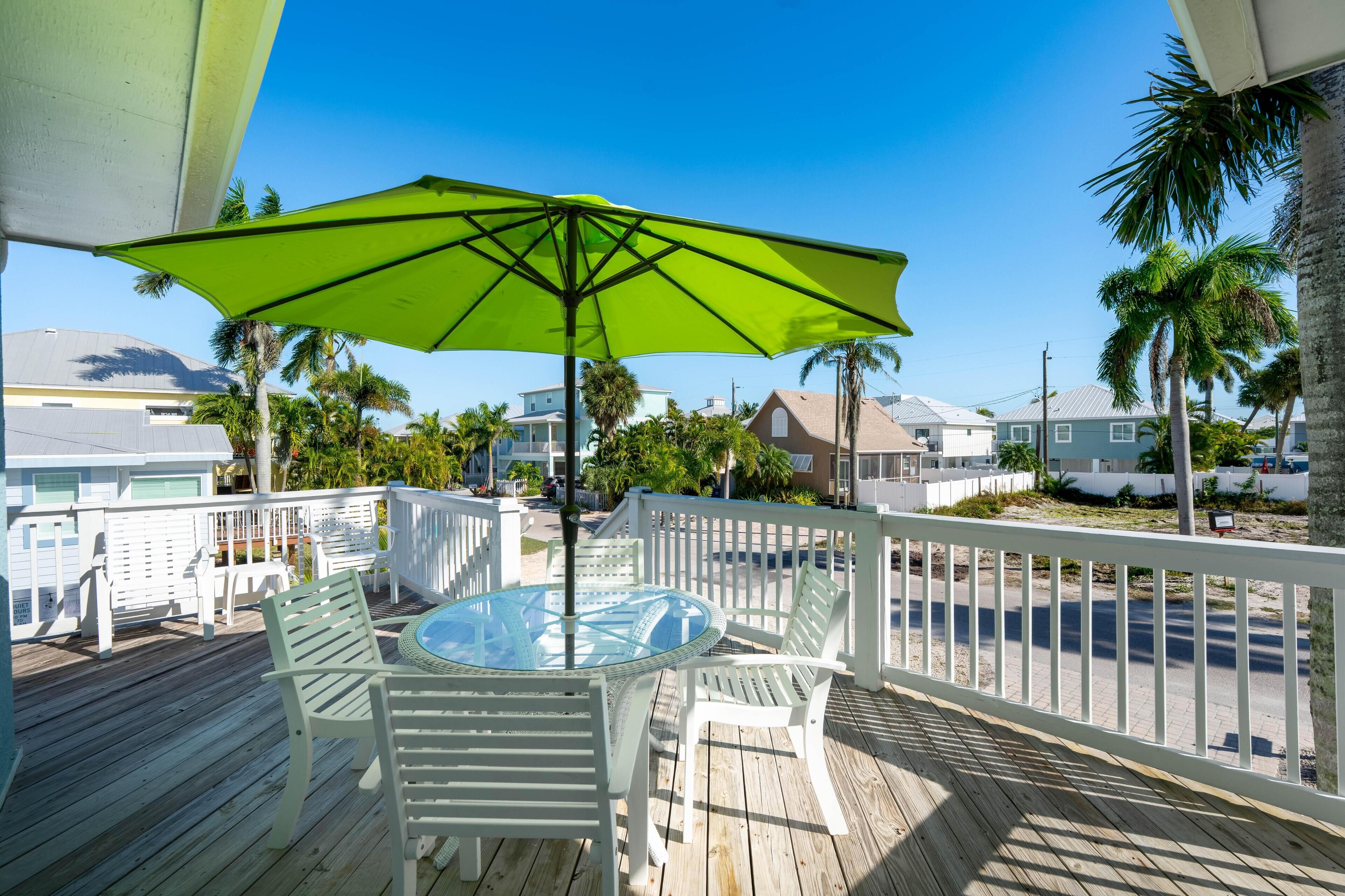 A wooden deck with a glass table and white chairs, a green umbrella, and a view of houses and palm trees.