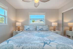A bedroom with a bed, two nightstands, two lamps, a window with a view of the beach, and a ceiling fan. - Blue Pearl Manor Upper (Fort Myers Beach)