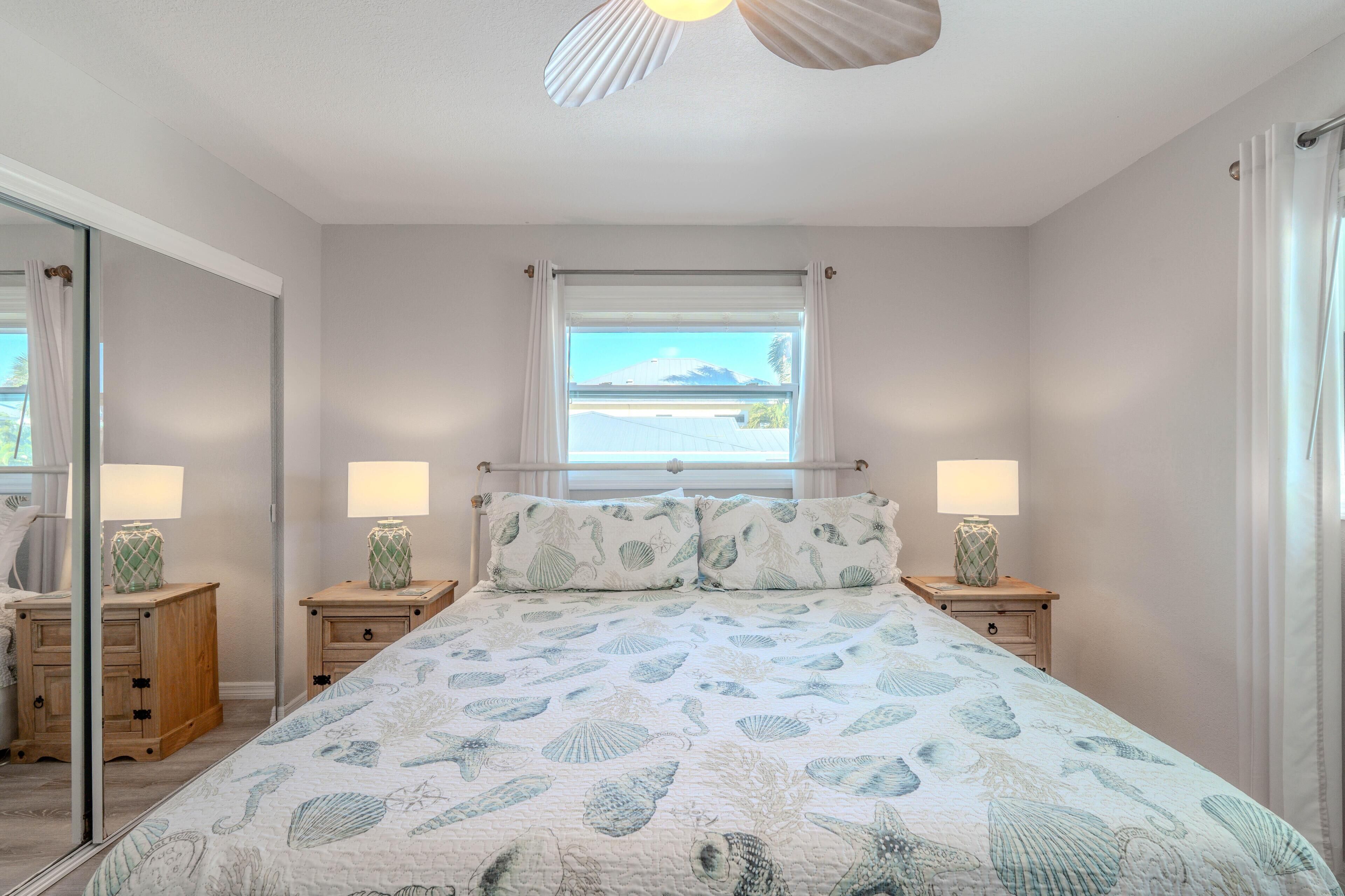 A bedroom with a bed, two bedside tables with lamps, a window with a view, and a ceiling fan.