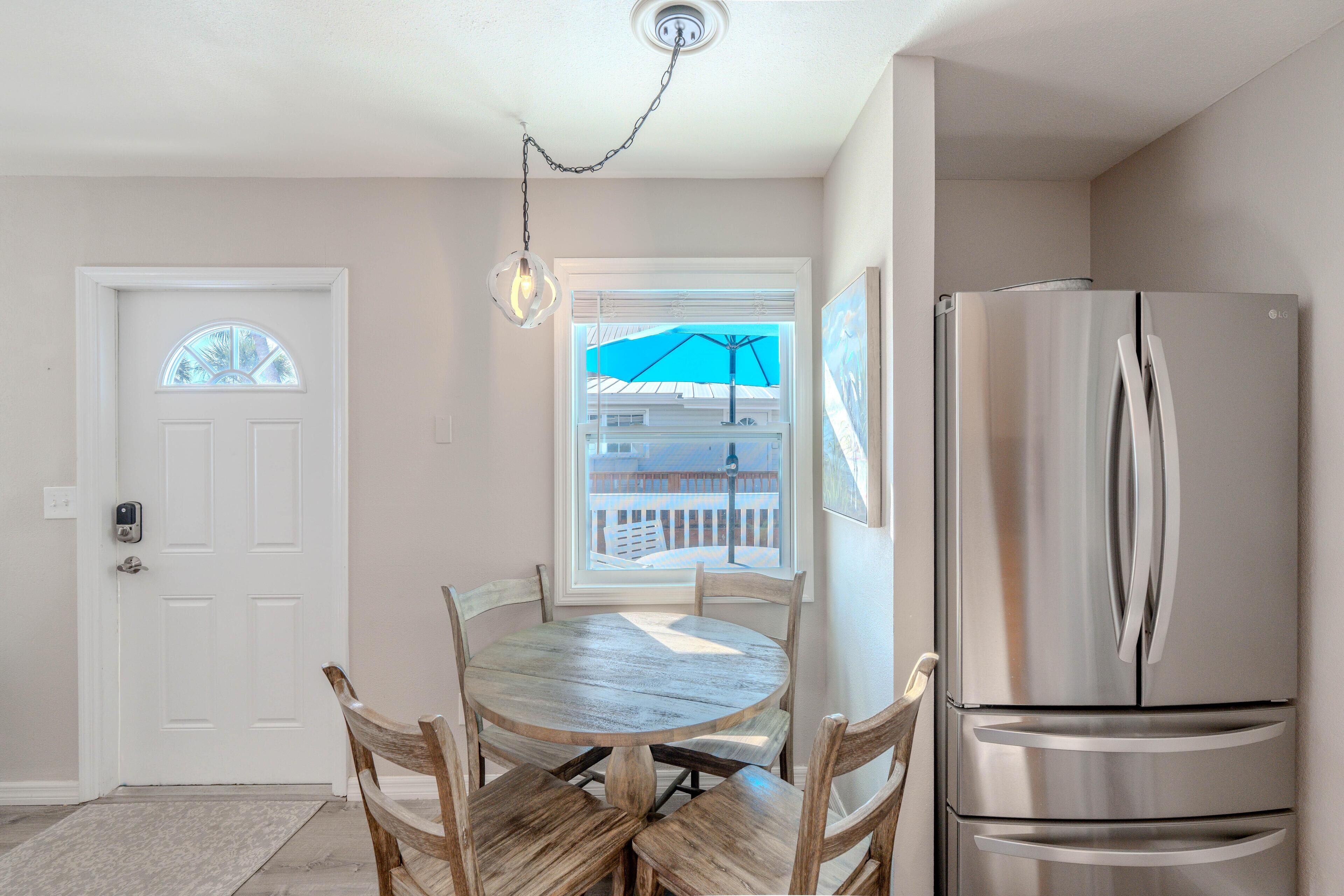 A kitchen with a stainless steel refrigerator, a wooden dining table with chairs, a white door, and a window with a view of a balcony.
