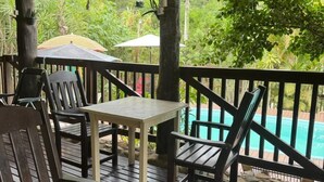 Un terrazza in legno con tavolo e sedie, affacciata su una piscina e una vegetazione rigogliosa.