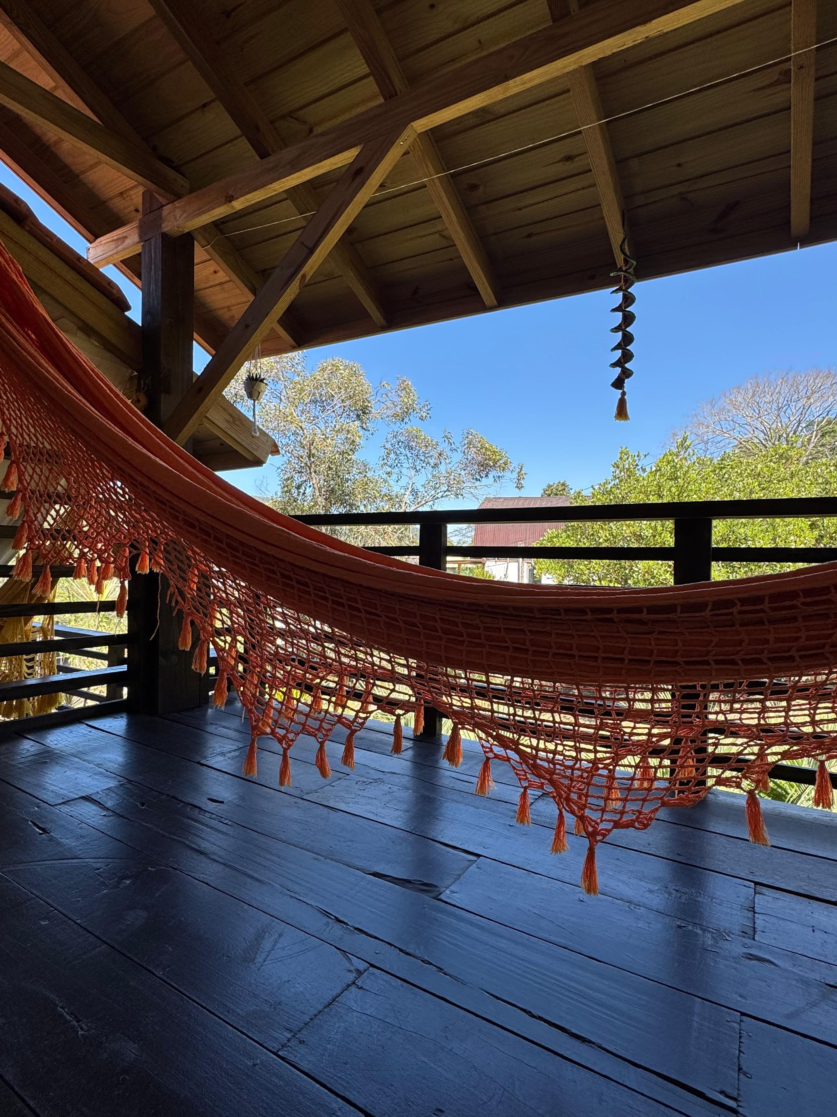 A hammock under a wooden structure with a view of trees and a building in the background.
