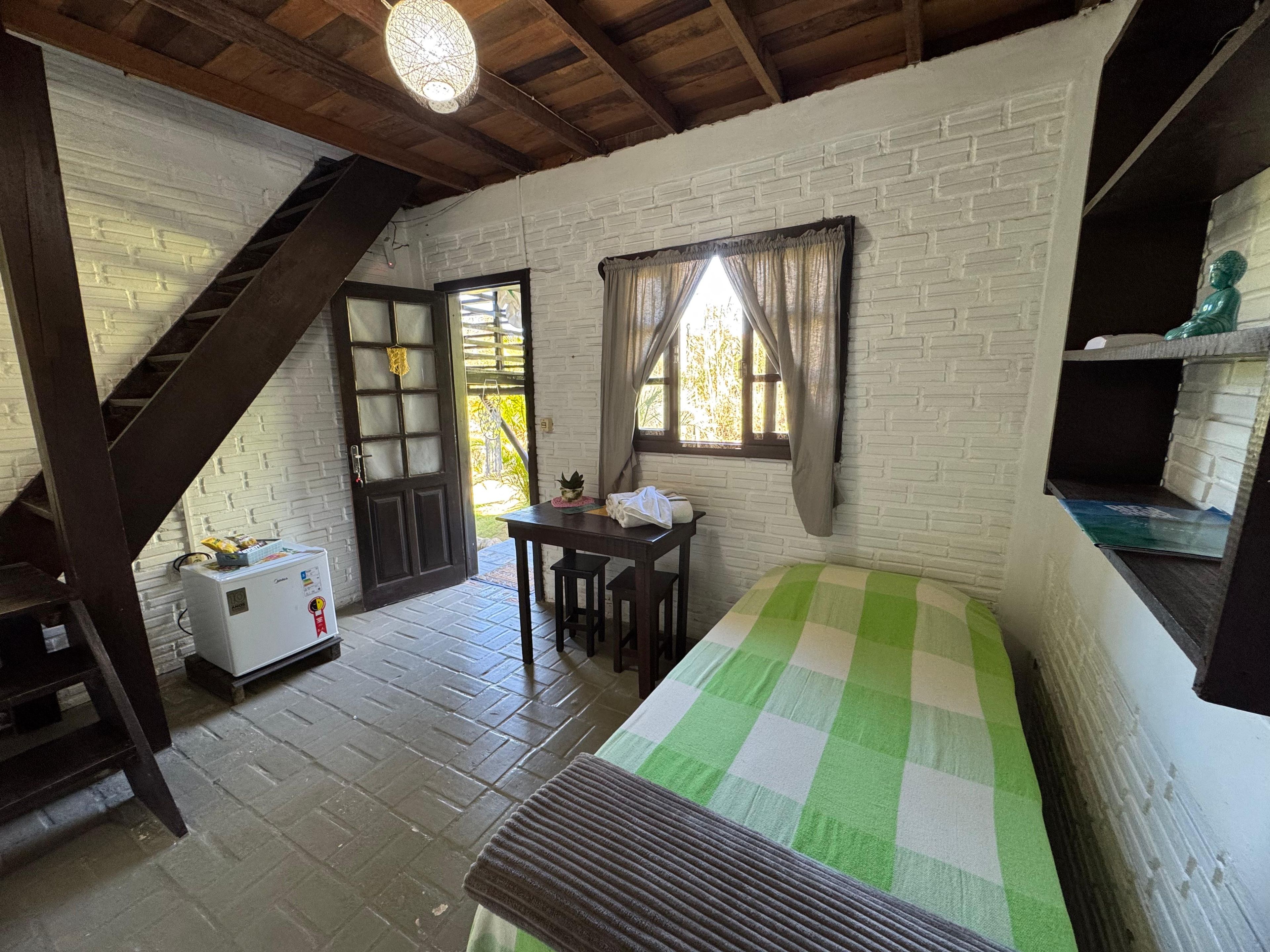 Una habitación rústica con una cama, una mesa, una silla, un refrigerador pequeño, una escalera y una ventana con cortinas.