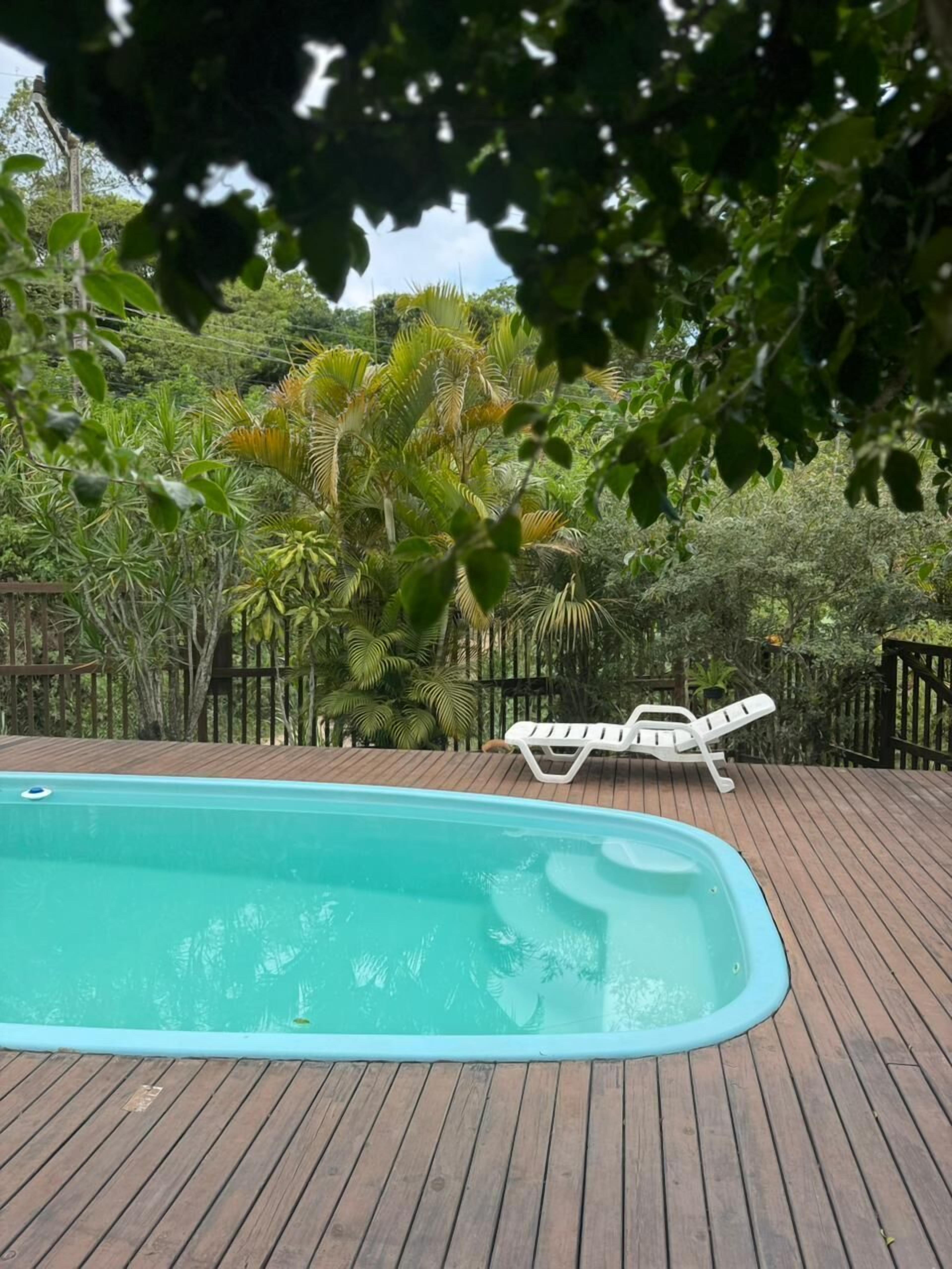 Um deck de madeira com piscina, cercado por vegetação exuberante e uma cadeira de lounge branca.