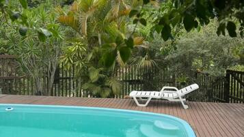 Um deck de madeira com piscina, cercado por vegetação exuberante e uma cadeira de lounge branca.