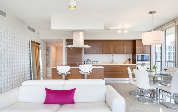 Un salon moderne avec un canapé blanc, une table à manger en verre et un coin cuisine doté de meubles en bois et d’appareils électroménagers en acier inoxydable.