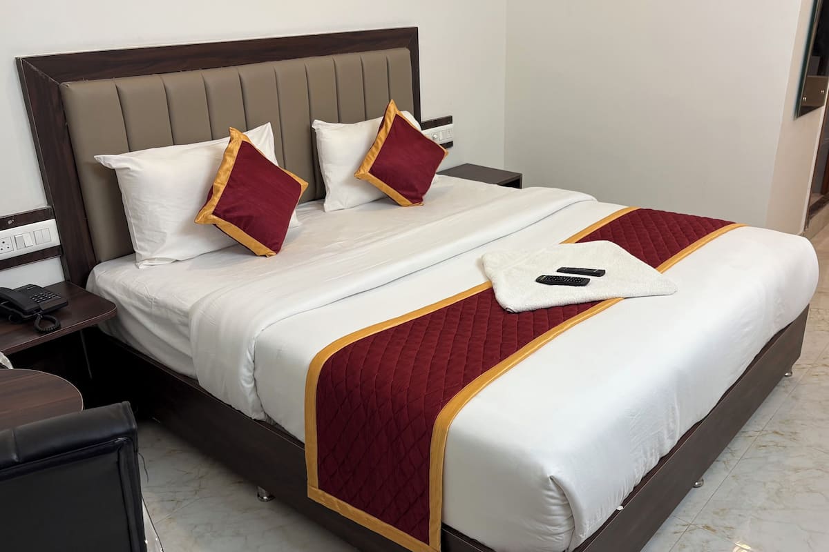 Una cama bien tendida con ropa blanca y granate, un detalle decorativo rojo acolchado y una toalla blanca con controles remotos.