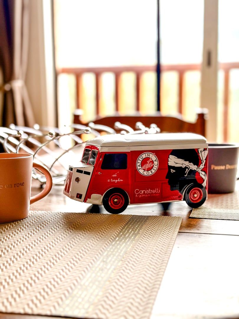Ein Spielzeug-Feuerwehrauto auf einem Tisch mit kariertem Tischläufer, einer Tasse und einem Stuhl im Hintergrund.
