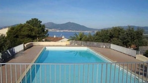 Ein rechteckiger Swimmingpool mit einem Metallzaun, umgeben von einer gepflasterten Terrasse. Im Hintergrund sind Bäume, ein Berg und ein Gewässer zu sehen.