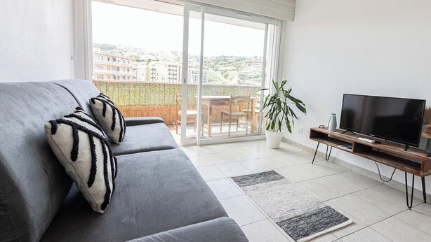 Uma sala de estar moderna com um sofá cinzento, uma TV de tela plana e uma porta de vidro deslizante que dá para uma varanda.