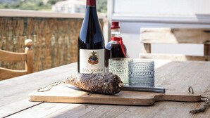 Uma mesa de madeira com uma garrafa de vinho San Michele, um copo e uma fatia de carne curada em uma tábua de corte.