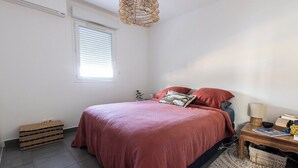 A bedroom with a bed, a bedside table with a lamp, a window with blinds, and a woven basket. - "I Sanguinari" – 2BR Sea View Apt | by Efficios (Ajaccio)