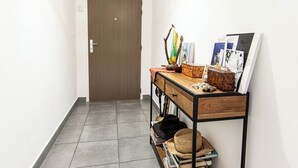 A hallway with a wooden door, a small wooden table with various items, and a wall-mounted shelf with hats and books. - "I Sanguinari" – 2BR Sea View Apt | by Efficios (Ajaccio)