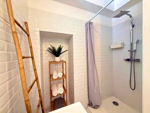 A shower with a handheld showerhead, a towel rack with towels, a wooden shelf with a plant, and a ladder against a white tiled wall. - "U Roccapina" – Downtown Studio | by Efficios (Sartène)