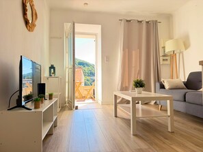 A living room with a TV, a table, a sofa, and a view of a building through an open door. - "U Roccapina" – Downtown Studio | by Efficios (Sartène)