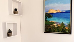 A framed painting of a coastal landscape, white wall, and two small white shelves with potted plants. - "A Punta Lisa" – Seaside 1BR | by Efficios (Ajaccio)