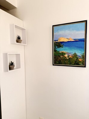 Un tableau encadré représentant un paysage côtier, un mur blanc et deux petites étagères blanches ornées de plantes en pot.