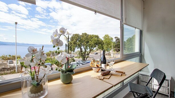 Ett modernt rum med ett stort fönster med utsikt över en vattenyta, ett träbord med en flaska, glas och snacks samt en stol.