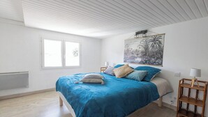 Un dormitorio con cama, ventana, radiador, un cuadro y una mesita de noche con lámpara.