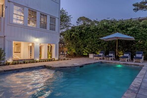 A swimming pool with lounge chairs and an umbrella, surrounded by a white building with large windows and a well-maintained garden.