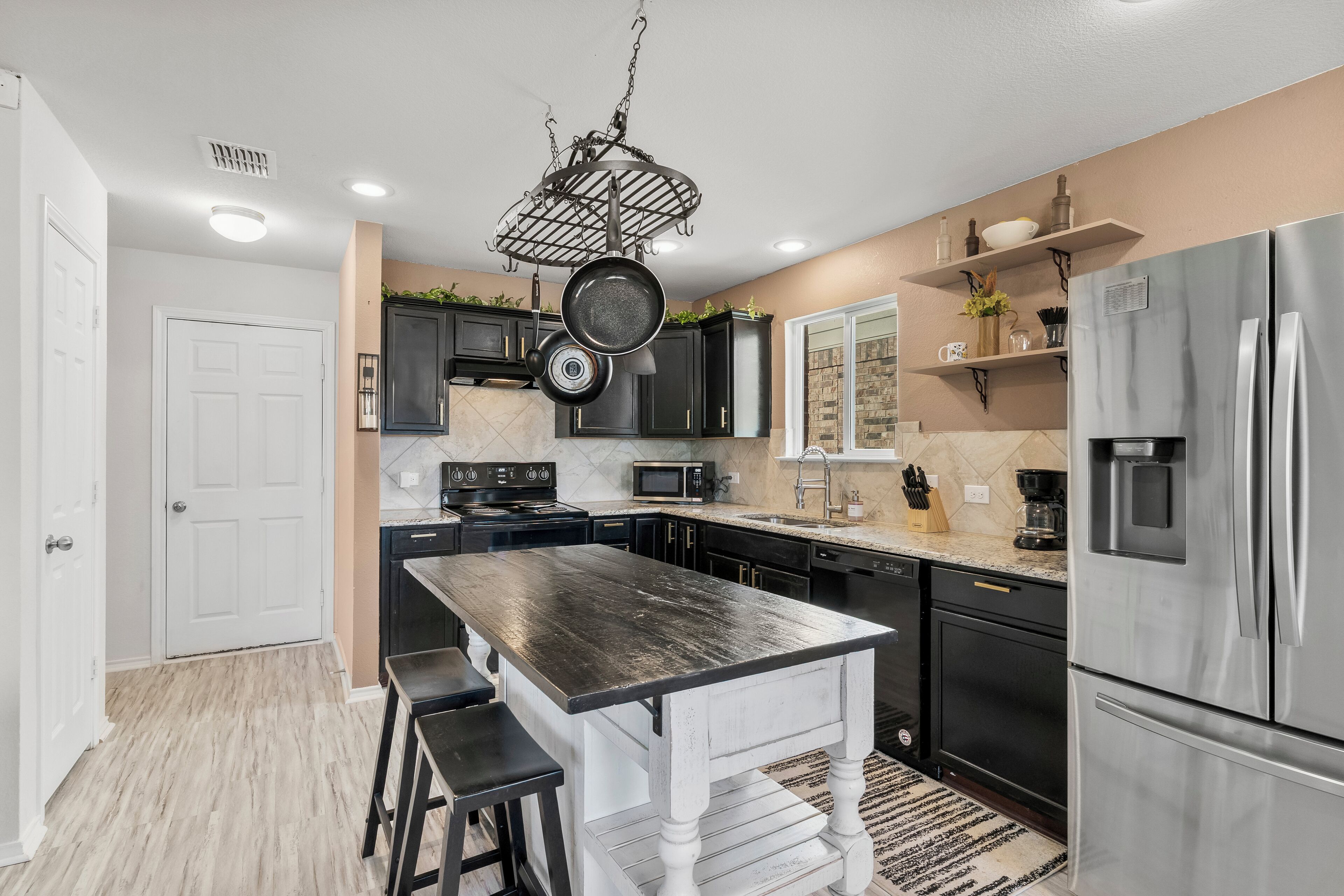 A modern kitchen with a central island, black cabinets, stainless steel appliances, and a hanging pot rack.