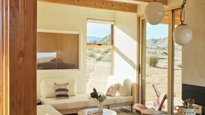 A modern living room with a sofa, coffee table, and dining table. Large windows offer a view of the desert landscape.