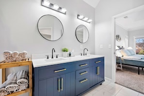 A modern bathroom with a dark blue vanity, two round mirrors, and a wooden shelf with towels. - Spacious 4BR Family Retreat Near Mall of GA & Lake Lanier (Sugar Hill)