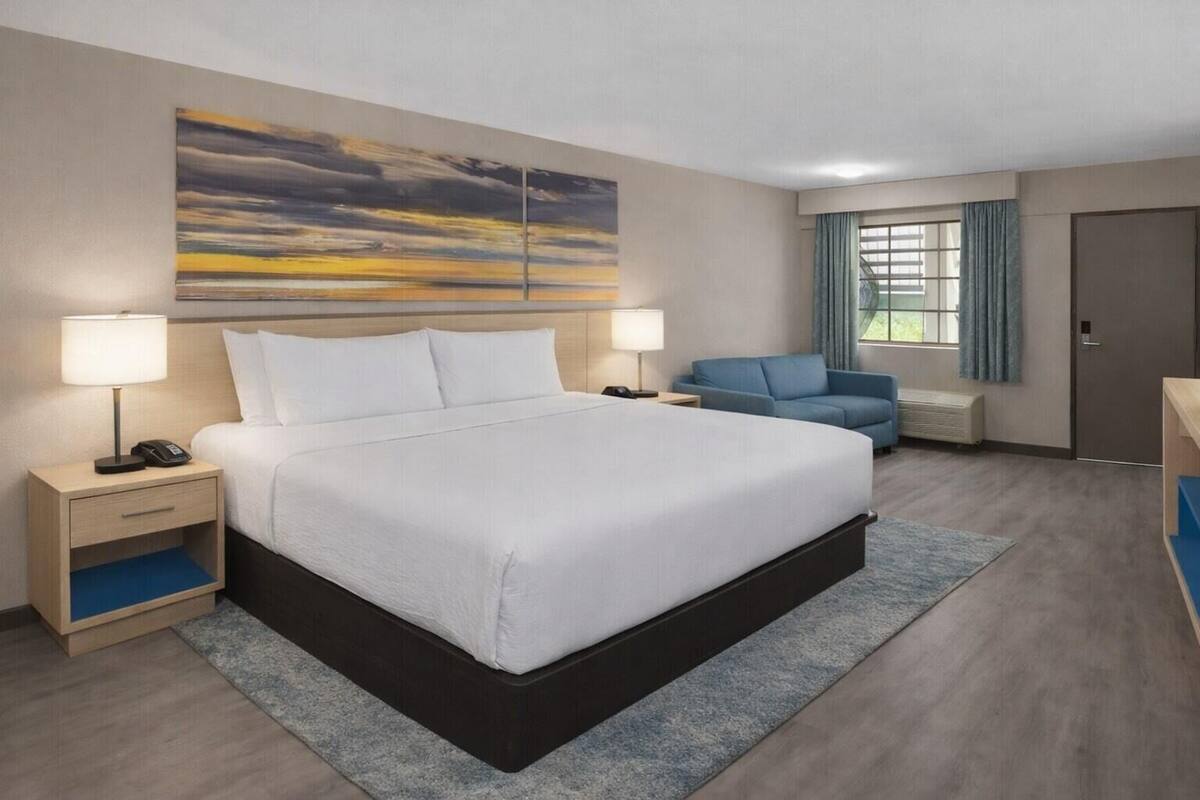 Een hotelkamer met een groot bed, twee nachtkastjes met lampen, een blauwe bank, een groot raam met gordijnen en een schilderij aan de muur.