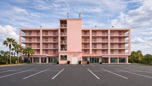 Ein pinkfarbenes, mehrstöckiges Hotel mit einem zentralen Turm, einem Parkplatz und Palmen.