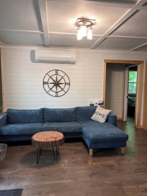Ein Wohnzimmer mit einem Ecksofa, einem runden Holztisch, einer Wanddekoration mit Kompass und einer Klimaanlage.