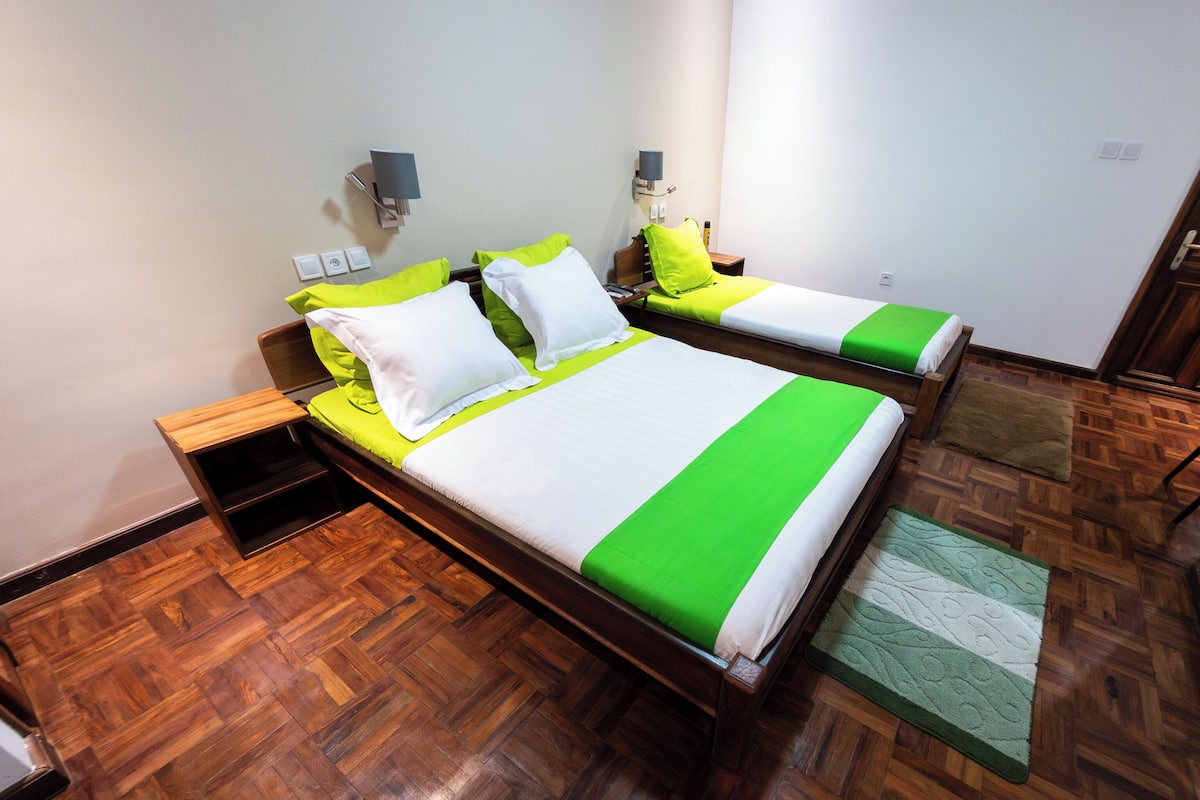 A double bed with green and white bedding, a bedside table, a nightstand, and a wall-mounted lamp.