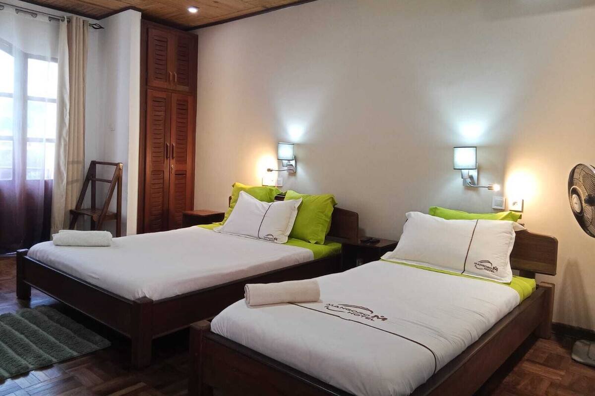 A hotel room with two beds, wooden headboards, a wooden ceiling, a green rug, a wooden chair, and a wall-mounted fan.