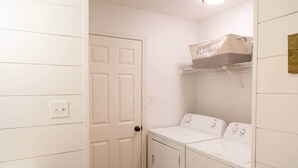 Un cuarto de lavandería con una puerta blanca, una lavadora, una secadora y un estante con una bolsa para ropa.