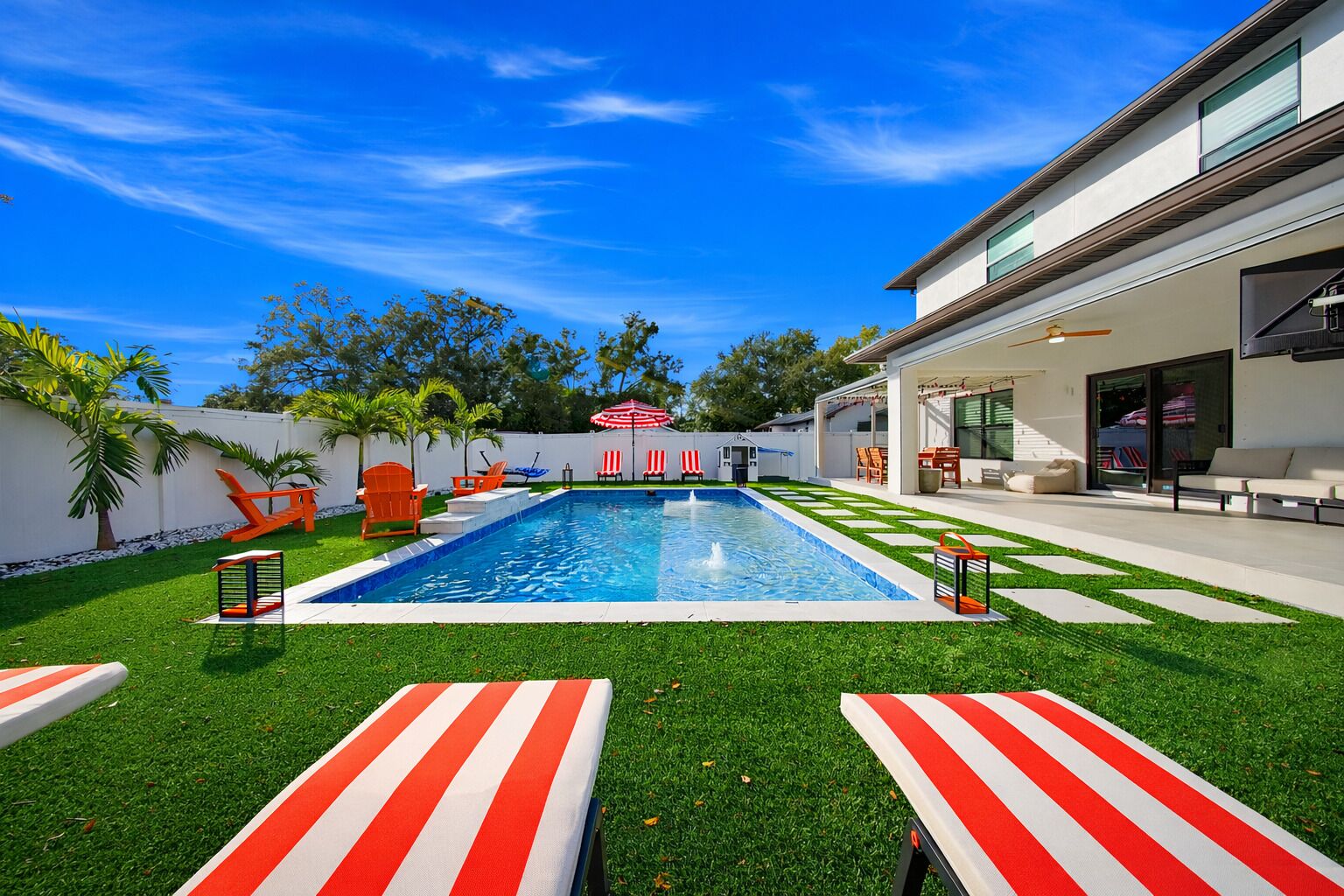A modern outdoor pool area with a clear blue sky, green artificial grass, and red and white striped lounge chairs.