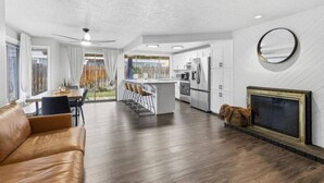 Una sala de estar moderna con un sofá, un comedor, una cocina con armarios blancos, una chimenea y vistas al exterior.