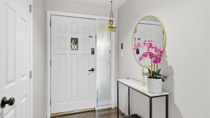 Un pasillo con una puerta blanca, una consola pequeña con un espejo y orquídeas rosadas, además de un banco.