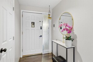 A hallway with a white door, a small console table with a mirror and pink orchids, and a bench.