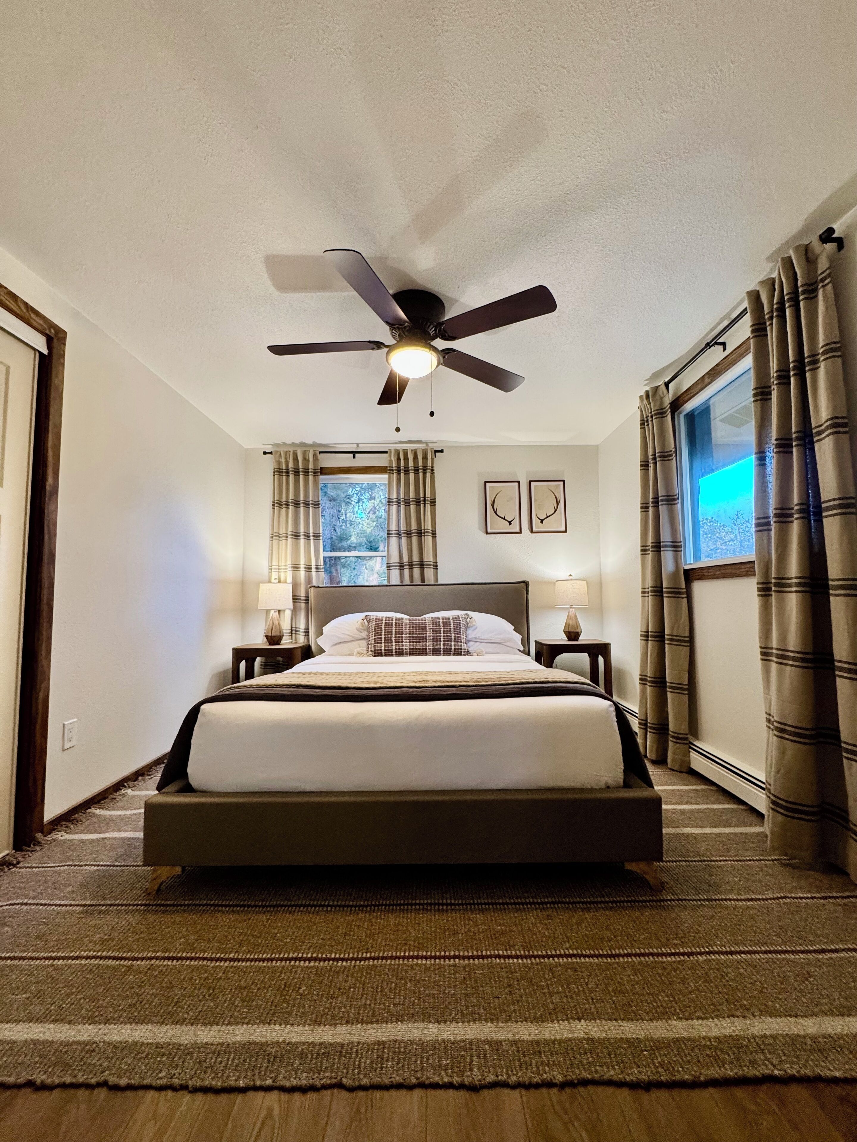 Un dormitorio con una cama, un ventilador de techo, dos mesitas de noche con lámparas, una ventana con cortinas e imágenes enmarcadas en la pared.