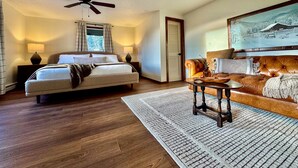 A bedroom with a bed, bedside lamps, a ceiling fan, a wooden floor, a rug, a leather sofa, and a painting on the wall.