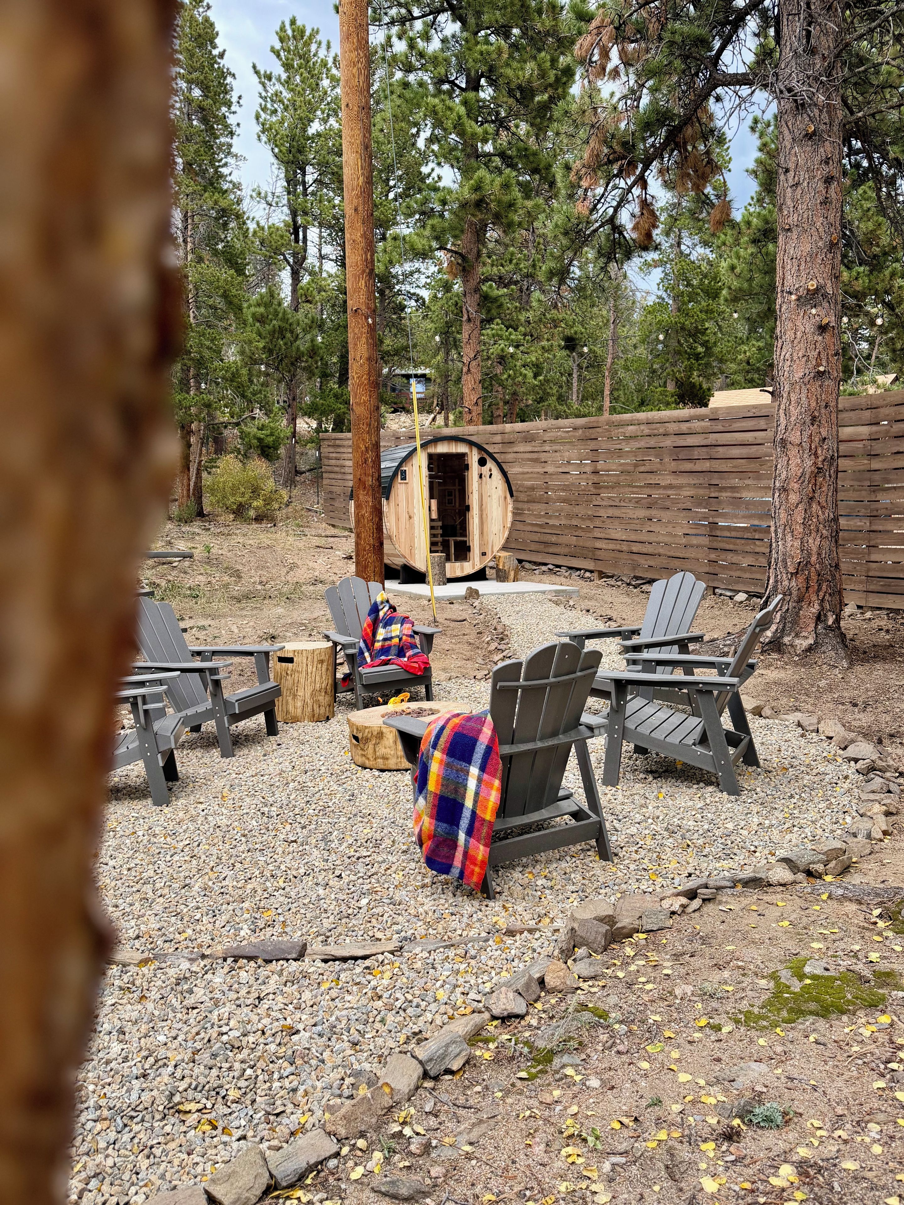 A rustic outdoor seating area with Adirondack chairs, a wooden barrel structure, and a gravel ground.