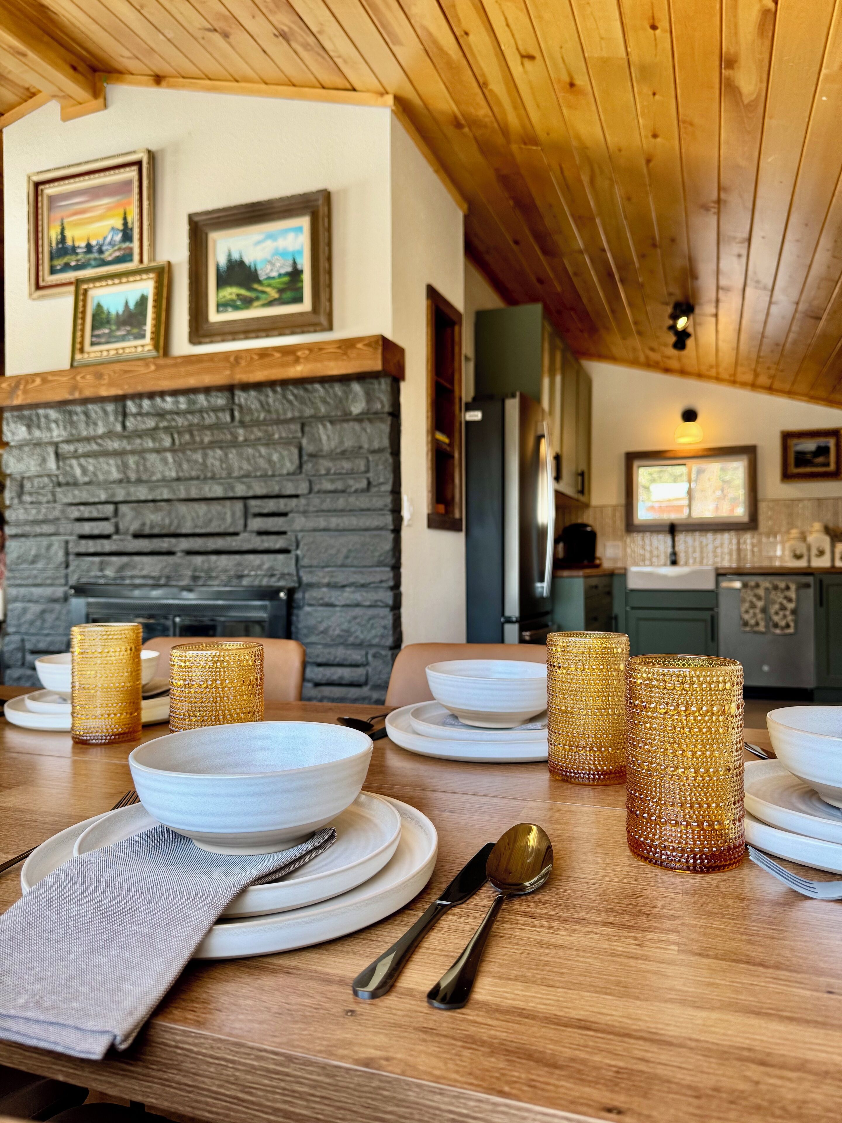 A dining area with a wooden table set for four, featuring white plates, bowls, and cutlery. A stone fireplace with framed pictures above it is visible.