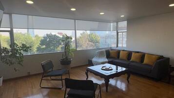 A spacious living room with a sofa, two chairs, a coffee table, and a large window offering a view of trees and buildings.