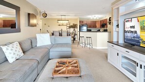 A modern living room with a grey sectional sofa, a wooden coffee table, and a kitchen area with wooden cabinets and a black refrigerator.