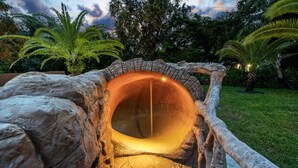 A stone tunnel with a warm light source inside, surrounded by rocks and palm trees. - One and Only Lazy River Pool Villa (North Miami)