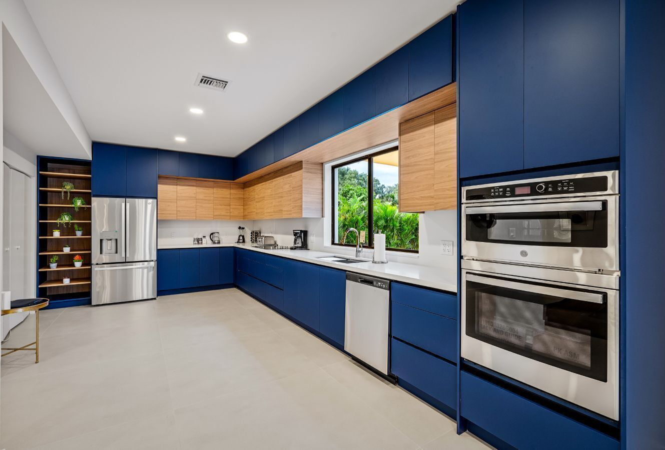 A modern kitchen with blue cabinetry, stainless steel appliances, and a window providing a view of greenery outside.