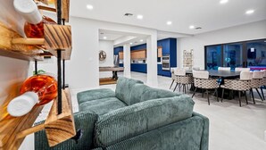A modern living room with a green sofa, wooden shelves with bottles, a dining area with a table and chairs, and a kitchen in the background. - One and Only Lazy River Pool Villa (North Miami)