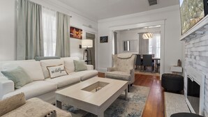 Een moderne woonkamer met een witte bank, een salontafel en een open haard. In de achtergrond staat een eethoek met een kroonluchter.