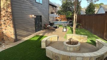 Un jardin avec un foyer en pierre, une piscine et un espace patio aménagé avec du mobilier d’extérieur.