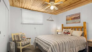 Un dormitorio con techo de madera, una cama con ropa de cama a rayas, una mecedora con cojines, un ventilador y un cartel motivacional.